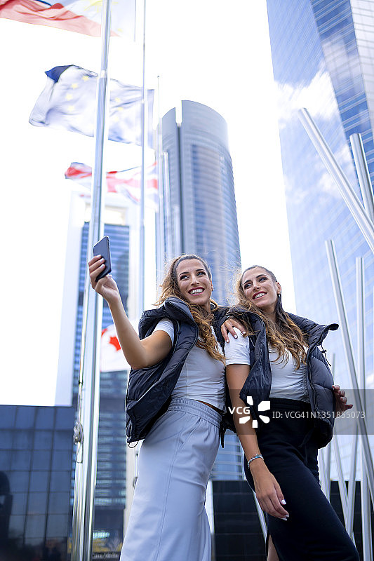 快乐的双胞胎姐妹在城市里用智能手机自拍图片素材