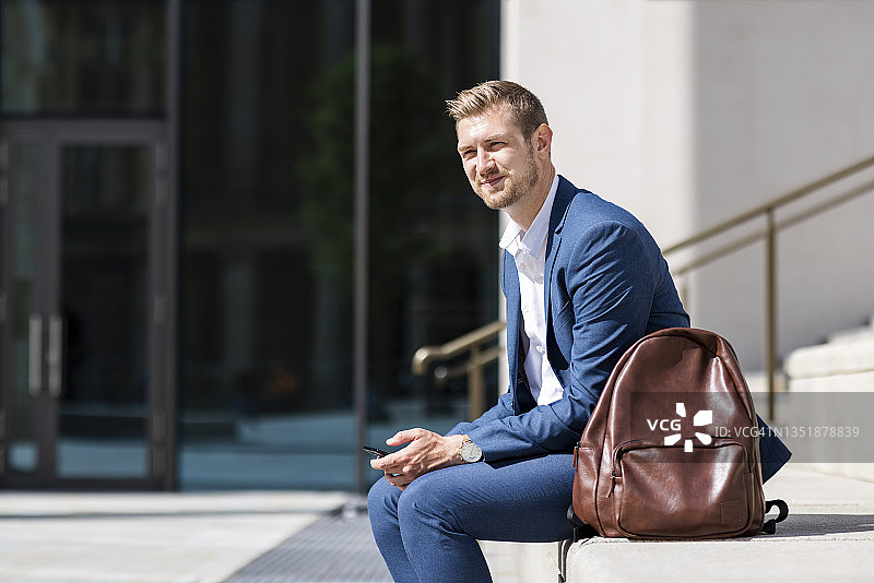坐在背包旁的微笑男商人，手持手机图片素材