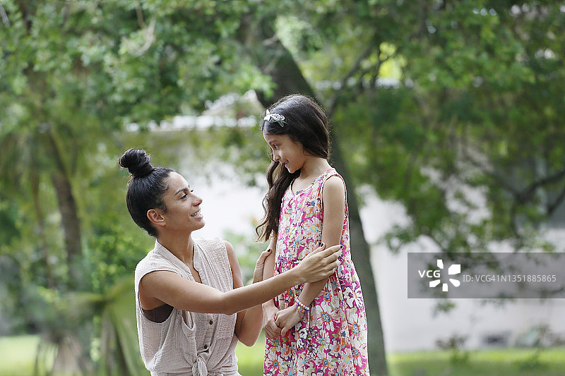 一位拉丁裔千禧一代女性跪下来与一个穿着飘逸连衣裙的黑发小女孩交谈，地点在绿树成荫的街道上，两人都穿着休闲服装。图片素材