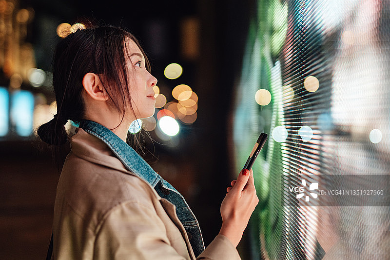 年轻女人在夜晚城市街道上对着LED屏幕使用手机，连接未来图片素材