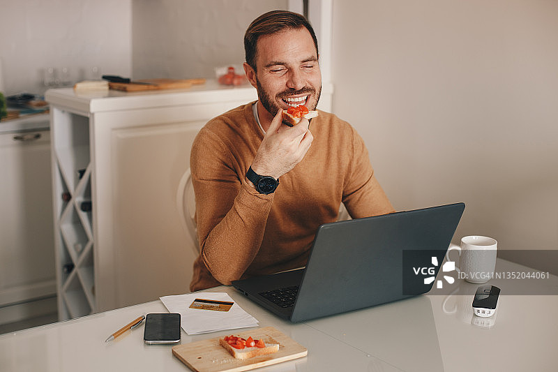 年轻自由职业者坐在桌旁吃三明治，面前放着笔记本电脑和咖啡杯。图片素材