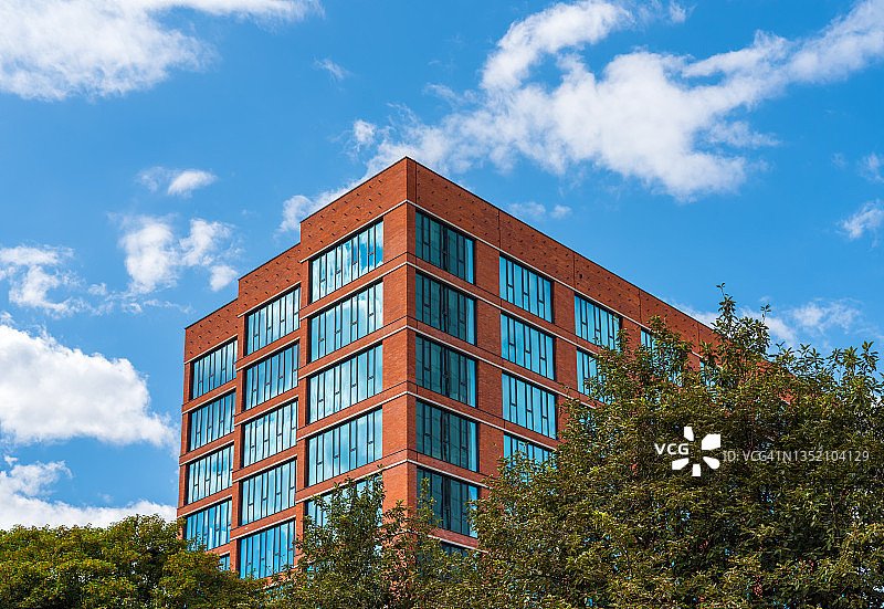 现代办公楼的外部仰视角度，具有红砖作品的立面和大窗户。前景有很多树木图片素材