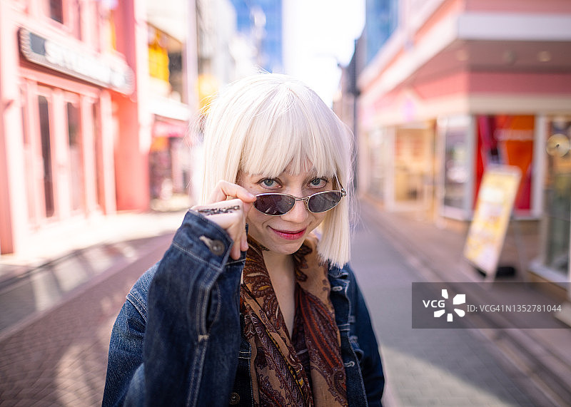 在粉色购物街上看镜头的铂金色头发、戴着太阳镜的年轻女人图片素材