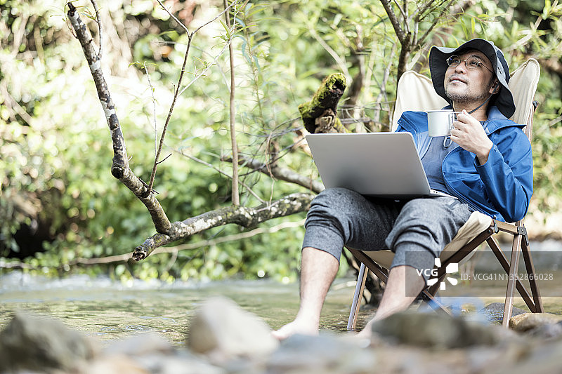 心理健康恢复与自然：老年男人在雨林溪流边坐在露营椅上喝咖啡和使用笔记本电脑放松图片素材