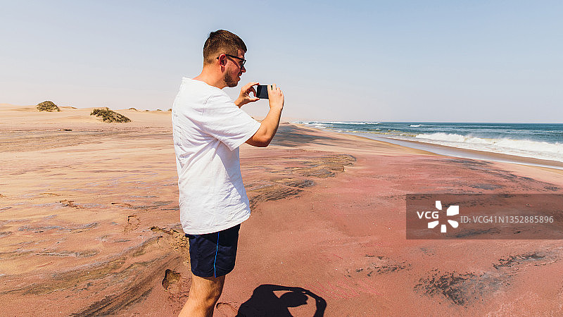 男子旅行者在纳米比亚海边用智能手机拍摄红色矿物沙图片素材