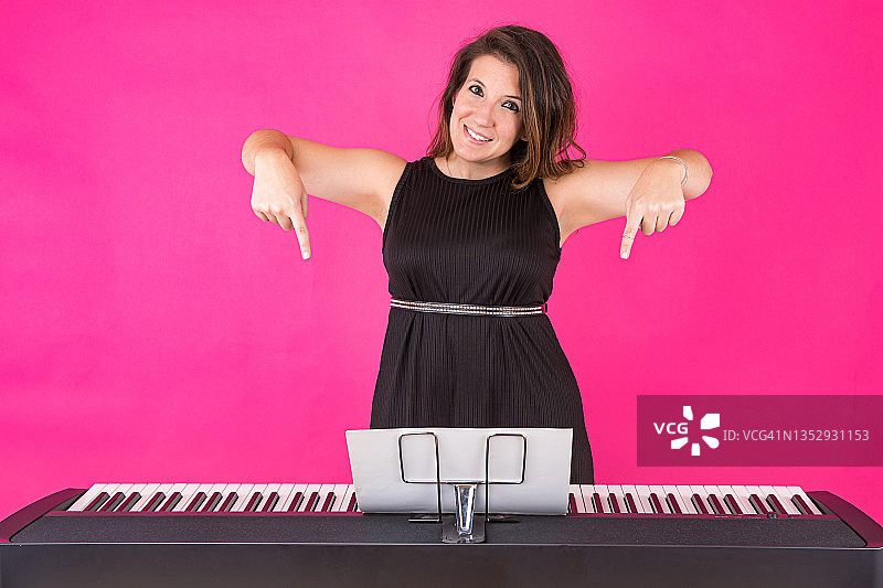 钢琴家女孩身穿黑色连衣裙，戴着高音谱号戒指，在粉红色背景上指向钢琴。音乐、钢琴家和作曲家的概念图片素材