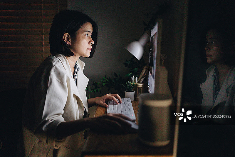 亚洲女人坐在办公桌前，用智能数码设备看着电脑，深夜研究项目。图片素材