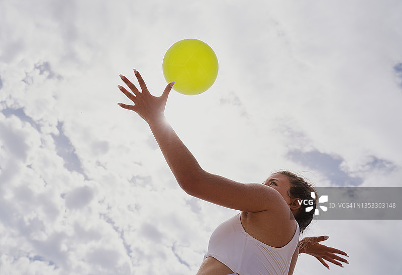白人女性打沙滩排球，专注地看着球，在多云的天空下发球。运动概念。图片素材