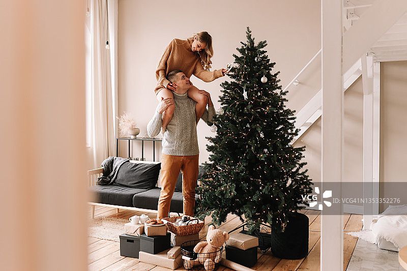 一对沉浸在爱河中的快乐情侣穿着毛衣，一起用气球和玩具装饰圣诞树，为舒适的客厅里的圣诞节做准备。男人和女人在新年的周末共度时光图片素材