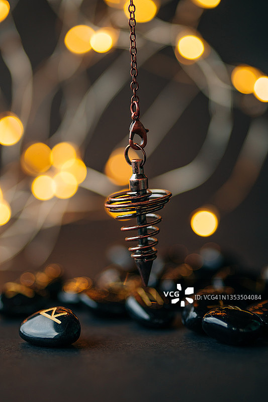 青铜探测摆锤和带有金色文字的黑曜石符文与花环灯背景。魔法深奥仪式概念。您的设计的复制空间图片素材