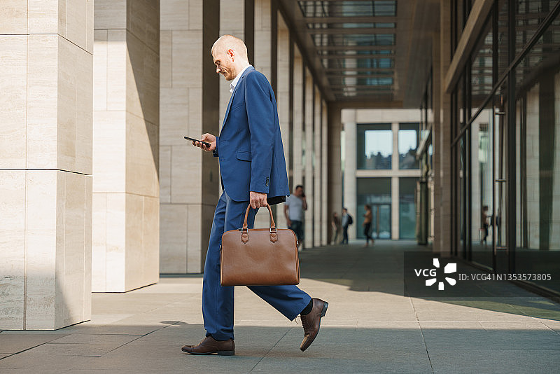 微笑的商人走在街上，使用智能手机并拿着公文包图片素材