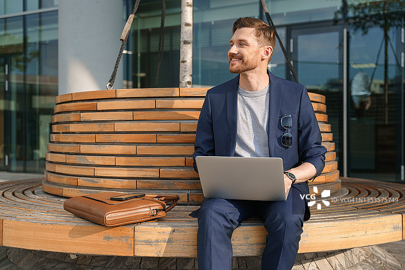 英俊的年轻商人身穿蓝色西装，坐在圆凳上看电脑，面带微笑图片素材