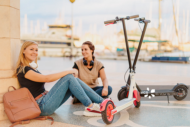 两位年轻女人和她们的电动滑板车一起坐在港口图片素材
