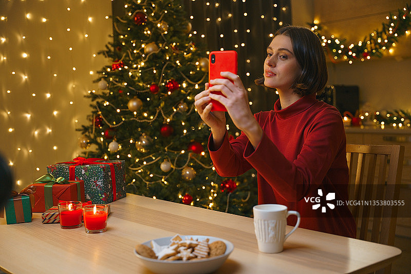 一位美丽的年轻女博主在圣诞主题内景中使用红色智能手机自拍图片素材