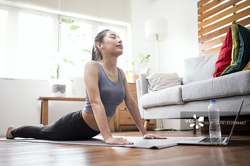 一位亚洲年轻女性在公寓的瑜伽垫上做眼镜蛇伸展运动，同时观看瑜伽虚拟教程。科技让家庭锻炼更轻松。图片素材