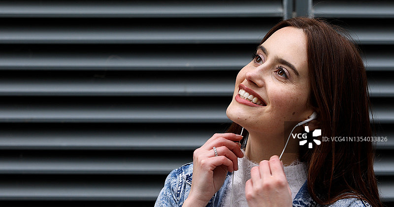 年轻女人戴着耳机听音乐，站在建筑物外的金属格栅前图片素材
