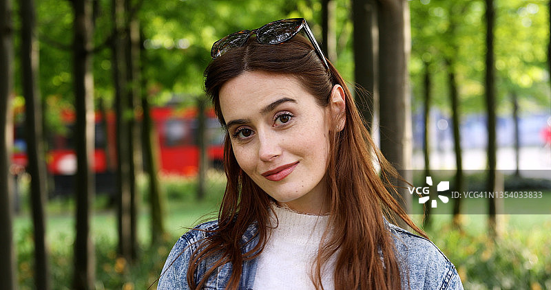 一位在城市公园里的年轻女子，头戴太阳镜，看着镜头微笑，远处有一辆红色伦敦巴士图片素材