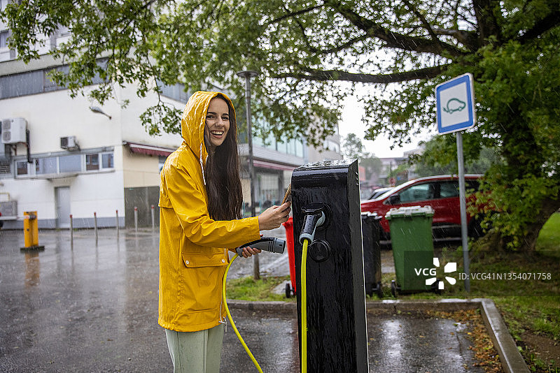 身穿雨衣的年轻女子用手机支付电动汽车充电费用图片素材