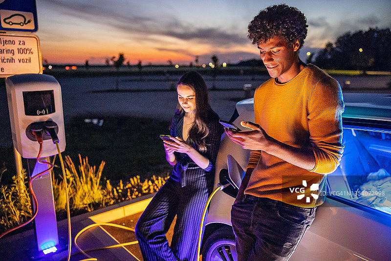 夫妇在使用手机，他们的电动汽车正在充电图片素材