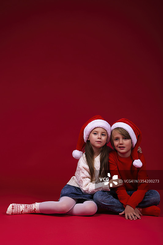一位穿着红色针织套头衫和蓝色牛仔裤以及圣诞红色袜子圣诞帽的可爱男孩，以及一位穿着白色套头衫和牛仔裙和白色紧身裤和圣诞帽的漂亮女孩的肖像图片素材