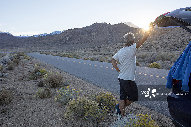 日出时分，男子在停在沙漠公路旁边的汽车附近放松图片素材