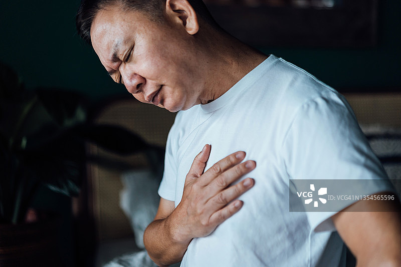 年长的亚洲男人胸口不适特写，坐在床上感到胸痛。老年人与健康问题概念图片素材