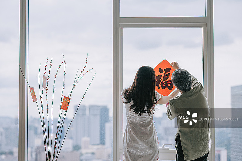 后视图中国新年妇女和她的女儿装饰房子的客厅与中国书法装饰的窗户准备家庭团聚图片素材