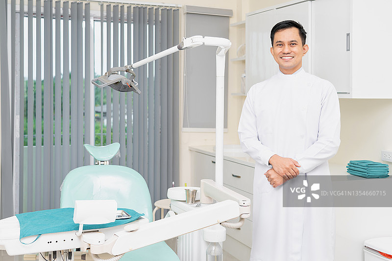 穿白色医生服的专业亚洲男牙医，带牙科椅和设备，面带微笑看着镜头图片素材