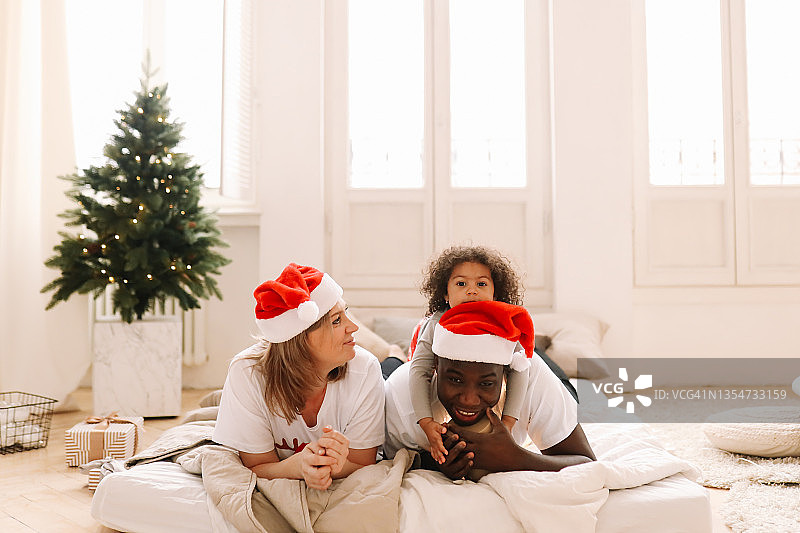 一个多种族混合家庭，带着一个戴着圣诞老人红色帽子的女儿，在舒适的卧室里装饰圣诞树，庆祝圣诞节图片素材