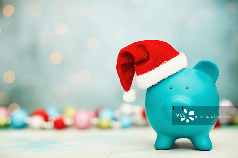 戴圣诞帽的蓝绿色小猪存钱罐与彩色圣诞装饰背景。圣诞储蓄或消费主题图片素材