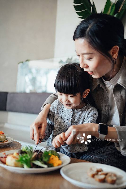 年轻的亚裔母亲和可爱的小女儿在咖啡馆里吃午饭，她们一起切着桌上的食物，一起享受快乐的一餐，家庭、食物和生活方式概念图片素材