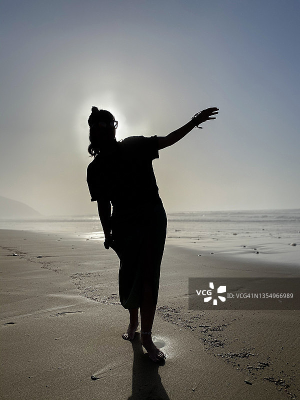 海滩上举起手臂的女人剪影竖版照片，在沙滩上行走，阳光在她身后图片素材