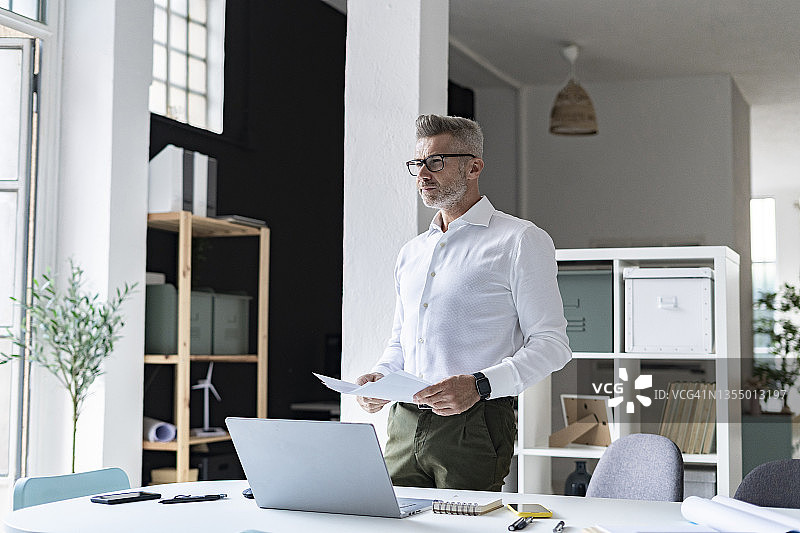 在办公室里，商人手持文件站在笔记本电脑前图片素材