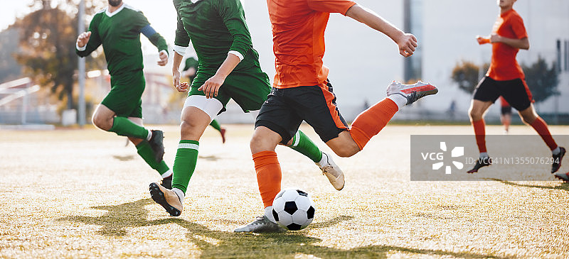 成年足球运动员在联赛中追逐并踢球。橙色和绿色球衣的足球运动员对抗图片素材
