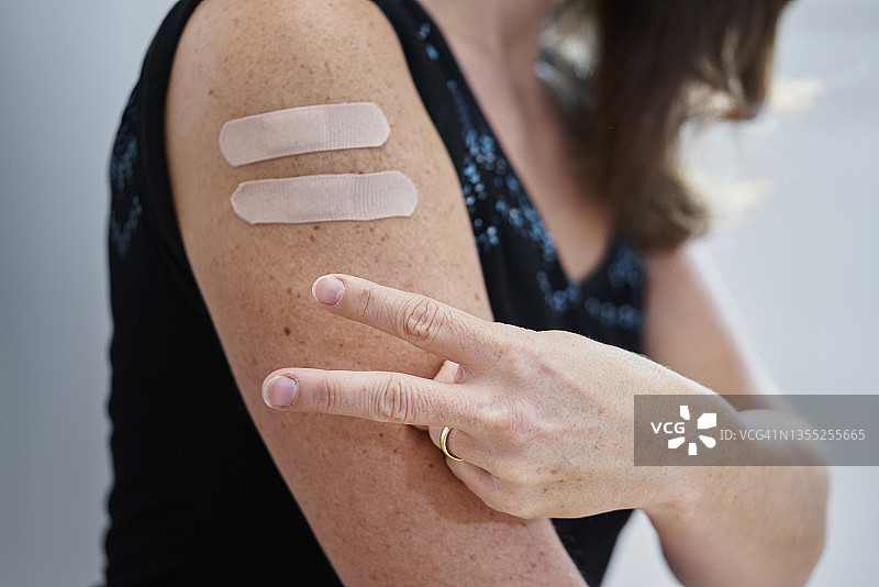 一名白人年轻女子手臂上贴着两个创可贴，用手指做出和平手势，等待针对模糊背景的第三剂疫苗的特写镜头图片素材