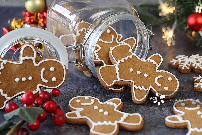 姜饼人饼干特写，用白色皇家糖霜装饰，从装满姜饼人的玻璃饼干罐中倾泻而出，迷你圣诞树，红色和金色小饰品，灰色背景，家庭烘焙图片素材