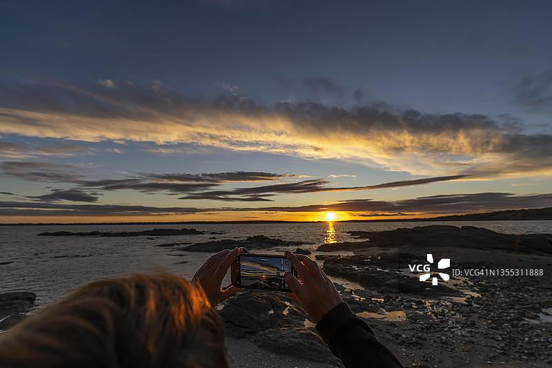 女人站在法国布列塔尼海岸的岩石上，用手机拍摄壮观的日落图片素材