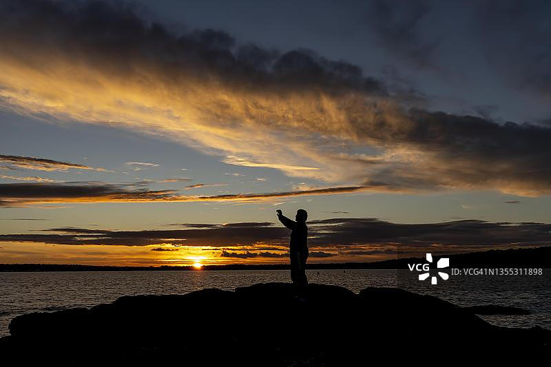 女人站在法国布列塔尼海岸的岩石上，用手机拍摄壮观的日落图片素材