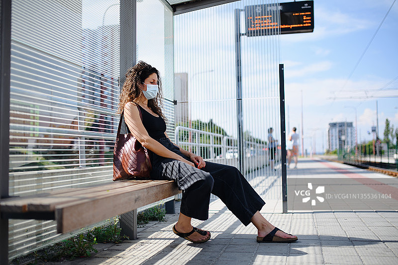 戴着口罩的中年妇女通勤者坐在电车站，在户外等候有轨电车的侧视图。新冠病毒概念图片素材