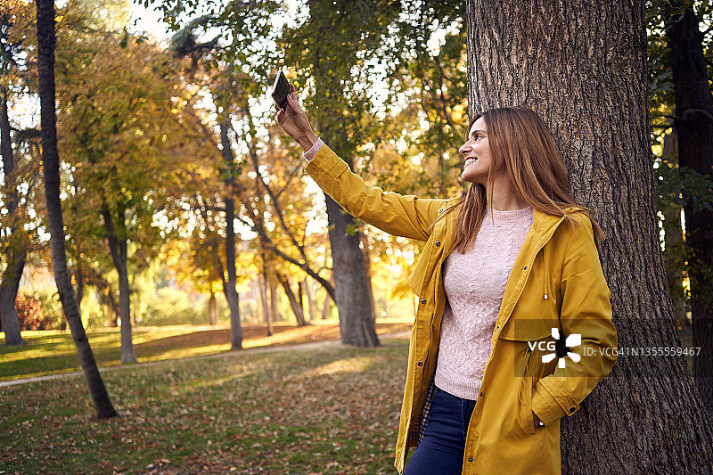 身穿休闲装和黄色外套的年轻女子靠在秋天公园的树干上，用手机进行视频通话图片素材