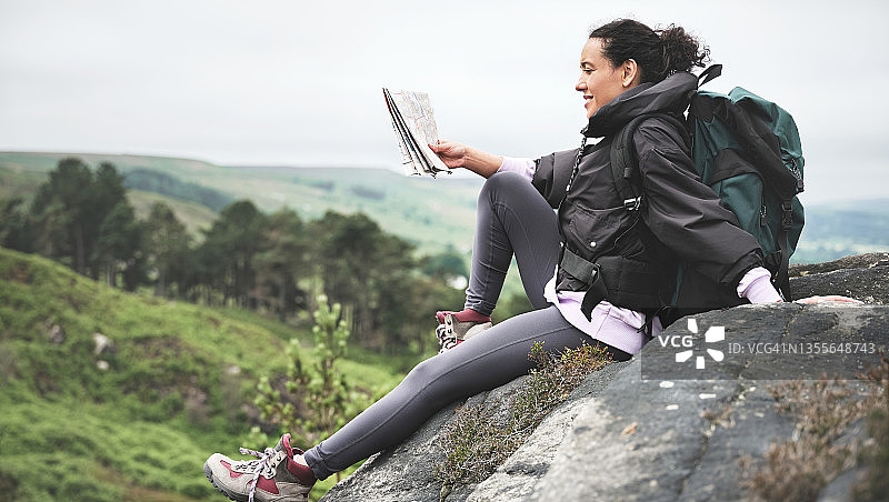一位迷人的年轻女子在徒步旅行中独自坐在岩石上阅读地图的全身照图片素材