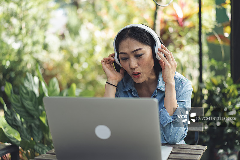 快乐的亚洲女性使用笔记本电脑，戴着耳机听音乐，同时在户外工作图片素材
