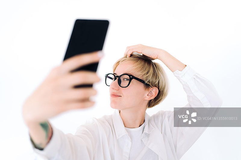 留着金色短发的年轻女子戴着眼镜整理发型，穿着白色休闲服装，性别中立概念图片素材