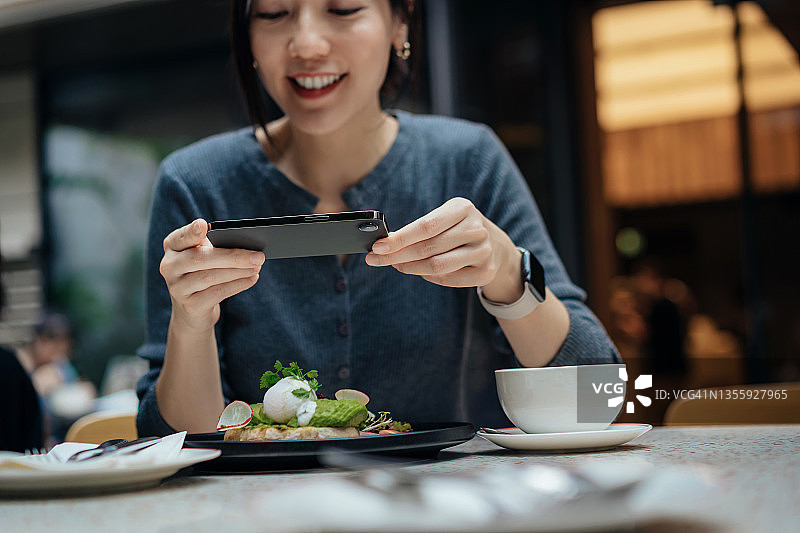 快乐的亚洲年轻女性在咖啡馆用智能手机拍摄美食并分享到社交媒体图片素材