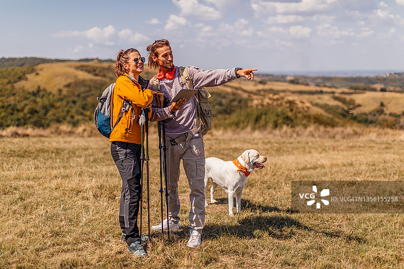 在秋日山脊上,两个年轻人和一只寻回犬坐在一起,拥抱他们的宠物,并背着徒步旅行的背包图片素材