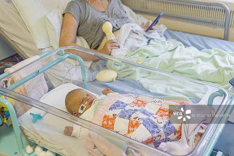 患有新生儿黄疸的女婴躺在医院婴儿床上，母亲在背景中表达母乳。图片素材
