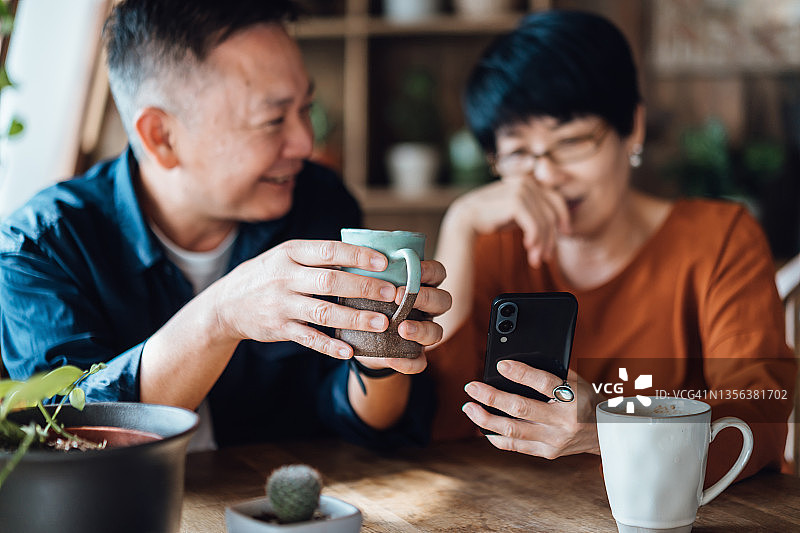 快乐的资深亚洲夫妇坐在咖啡馆，聊天，一起使用智能手机。享受放松的时刻。生活方式。老年人和技术图片素材