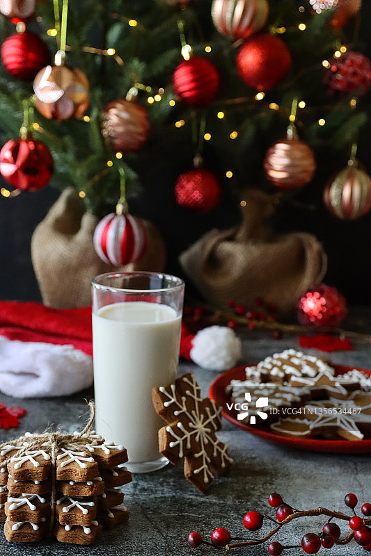 一堆糖霜姜饼雪花饼干用绳子系在一起的图像，饼干靠在圣诞主题饼干红盘旁边的牛奶玻璃杯上，圣诞老人的帽子，云杉圣诞树，粗麻布包裹的底座，红色和金色的小饰品，灰色背景图片素材