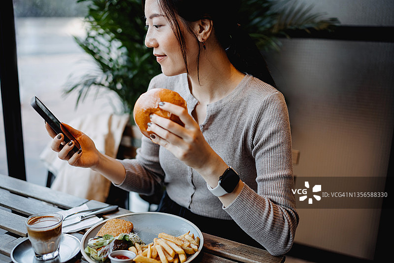 微笑的亚洲年轻女性在咖啡馆靠窗而坐，用智能手机吃午餐图片素材