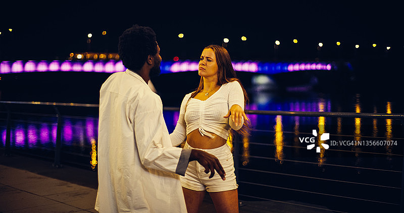 年轻情侣在河边大道上表演现代快节奏舞蹈，笑容满面，水面倒映灯光图片素材